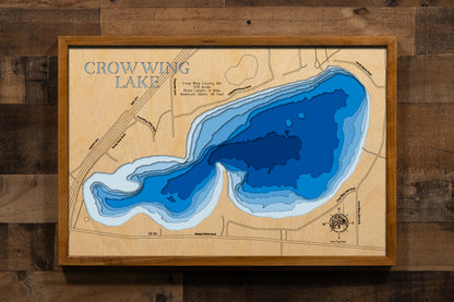A meticulously detailed, laser-cut, multi-layered wooden topographical map of Crow Wing Lake in Crow Wing County, Minnesota, showcasing the 379-acre shallow, fertile lake with depth contours from the 11-foot average depth to the 26-foot maximum. The natural wood top layer frames four miles of shoreline with aquatic vegetation and the public boat launch, while graduated blue layers create a gentle visual depth gradient.