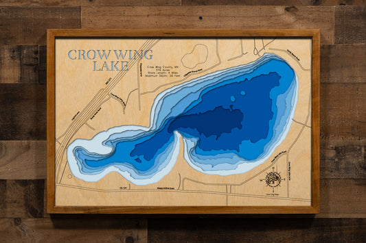 A meticulously detailed, laser-cut, multi-layered wooden topographical map of Crow Wing Lake in Crow Wing County, Minnesota, showcasing the 379-acre shallow, fertile lake with depth contours from the 11-foot average depth to the 26-foot maximum. The natural wood top layer frames four miles of shoreline with aquatic vegetation and the public boat launch, while graduated blue layers create a gentle visual depth gradient.
