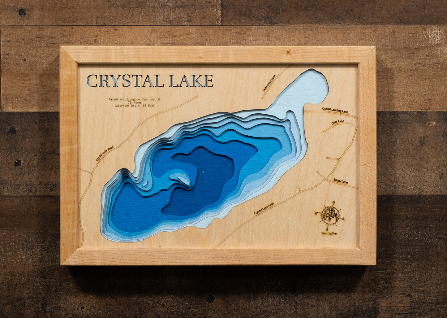 A laser-cut, layered wooden map of Crystal Lake in Forest and Langlade Counties, Wisconsin. The distinct shape of this natural lake, with its 75-acre surface, is clearly visible within the forested landscape of northern Wisconsin. The varying depths of the lake, reaching a maximum of 36 feet, are represented by different layers of wood, illustrating its underwater topography.