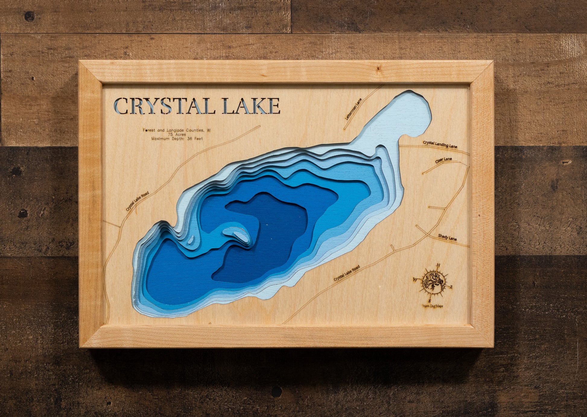 A laser-cut, layered wooden map of Crystal Lake in Forest and Langlade Counties, Wisconsin. The distinct shape of this natural lake, with its 75-acre surface, is clearly visible within the forested landscape of northern Wisconsin. The varying depths of the lake, reaching a maximum of 36 feet, are represented by different layers of wood, illustrating its underwater topography.