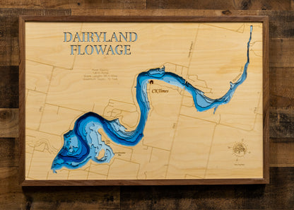 A framed wooden wall art piece titled "DAIRYLAND FLOWAGE" showing a stylized blue river or lake map etched onto a light wood background, mounted on a dark wood plank wall.