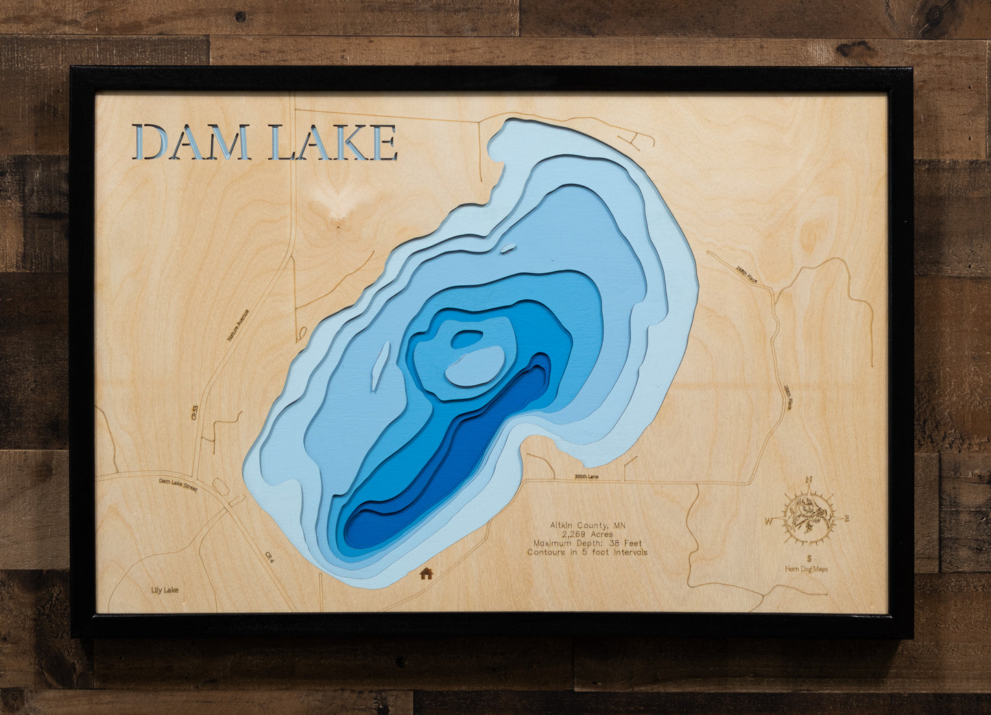 A laser-cut, layered wooden map of Dam Lake in Aitkin County, Minnesota. The shape of the lake is clearly defined within the northern Minnesota landscape. The varying depths of the lake, reaching a maximum of 38 feet, are represented by different layers of wood, with contour lines indicating depth changes at 5-foot intervals.