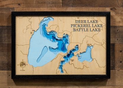 A precision laser-cut, multi-layered wooden map of the Deer, Pickerel, and Battle Lake chain in Itasca County, Minnesota. The natural wood top layer captures the interconnected shorelines of all three lakes — including Deer Lake's 25 islands — in fine detail, while a light-to-dark blue gradient highlights their dramatically varying depths. A distinctive, rustic statement piece for any Minnesota Northwoods cabin or lake home.