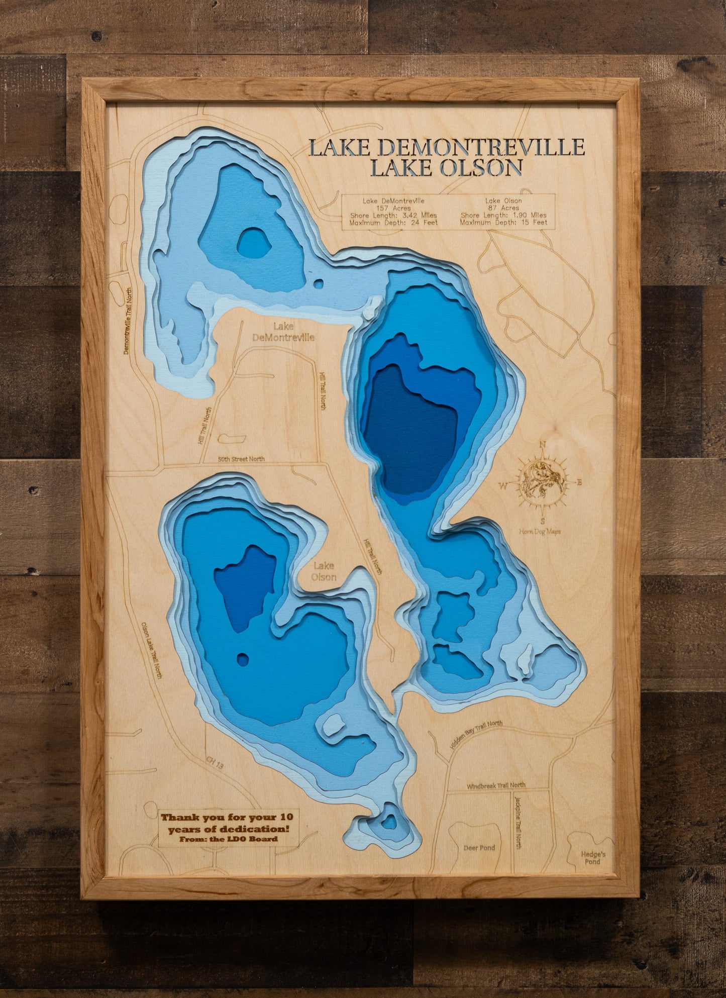 A laser-cut, layered wooden map depicting Lake DeMontreville and Lake Olson side-by-side in Washington County, Minnesota. The distinct shapes of both natural lakes are clearly visible within the suburban-natural landscape. The varying depths of Lake DeMontreville (maximum 24 feet) and Lake Olson (maximum 15 feet) are represented by different layers of wood, illustrating their individual underwater topographies.