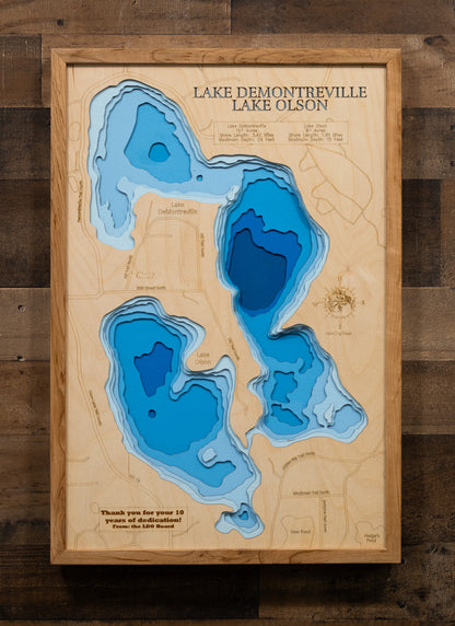 A laser-cut, layered wooden map depicting Lake DeMontreville and Lake Olson side-by-side in Washington County, Minnesota. The distinct shapes of both natural lakes are clearly visible within the suburban-natural landscape. The varying depths of Lake DeMontreville (maximum 24 feet) and Lake Olson (maximum 15 feet) are represented by different layers of wood, illustrating their individual underwater topographies.