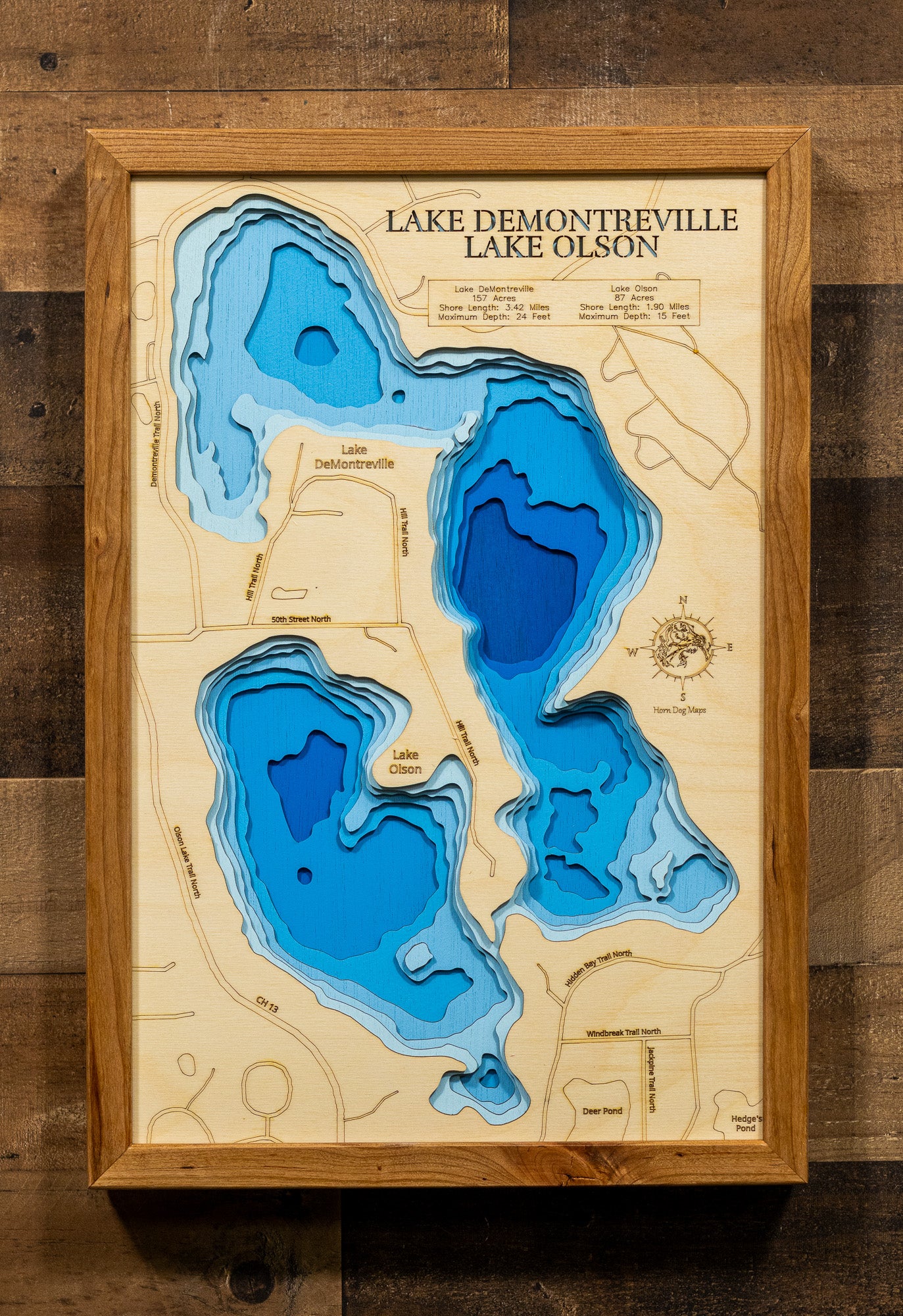 A laser-cut, layered wooden map depicting Lake DeMontreville and Lake Olson side-by-side in Washington County, Minnesota. The distinct shapes of both natural lakes are clearly visible within the suburban-natural landscape. The varying depths of Lake DeMontreville (maximum 24 feet) and Lake Olson (maximum 15 feet) are represented by different layers of wood, illustrating their individual underwater topographies.