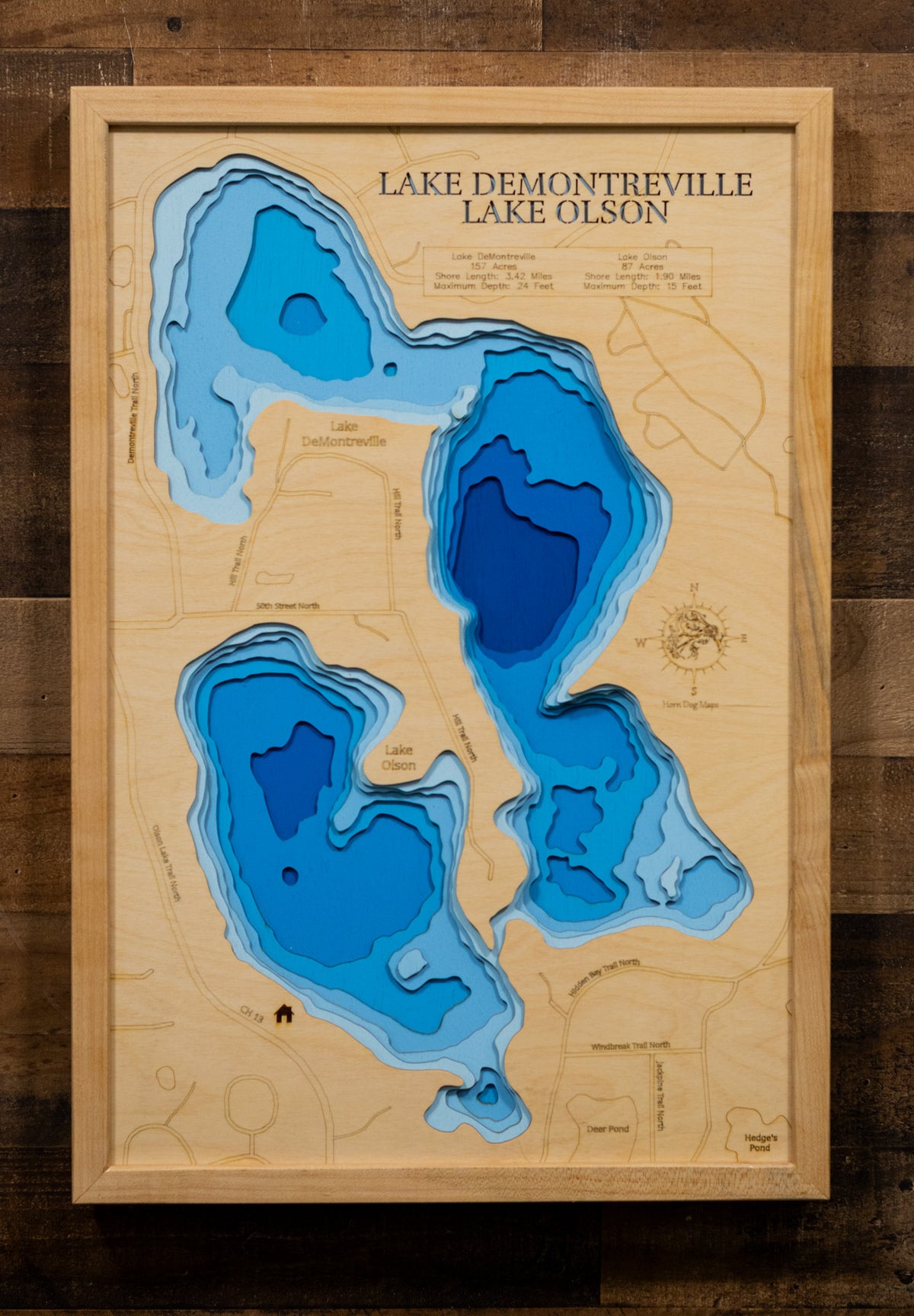 A laser-cut, layered wooden map depicting Lake DeMontreville and Lake Olson side-by-side in Washington County, Minnesota. The distinct shapes of both natural lakes are clearly visible within the suburban-natural landscape. The varying depths of Lake DeMontreville (maximum 24 feet) and Lake Olson (maximum 15 feet) are represented by different layers of wood, illustrating their individual underwater topographies.