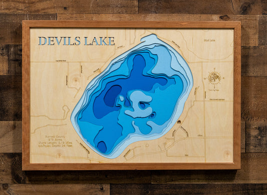 A framed wooden depth map of Devils Lake, featuring layered blue and cyan water contours on a light natural wood background, mounted on a dark wood plank wall.