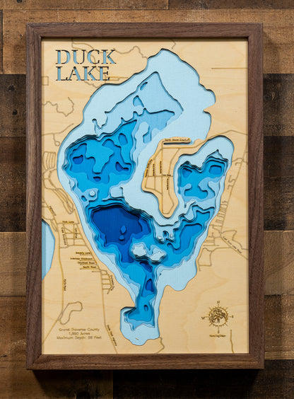 A laser-cut, layered wooden map of Duck Lake in Grand Traverse County, Michigan. The distinct shape of this 1,930-acre lake is visible within the northwestern Lower Michigan landscape. The varying depths of the lake, reaching a maximum of 98 feet and detailed by 10-foot contour intervals, are represented by different layers of wood, illustrating its underwater topography.