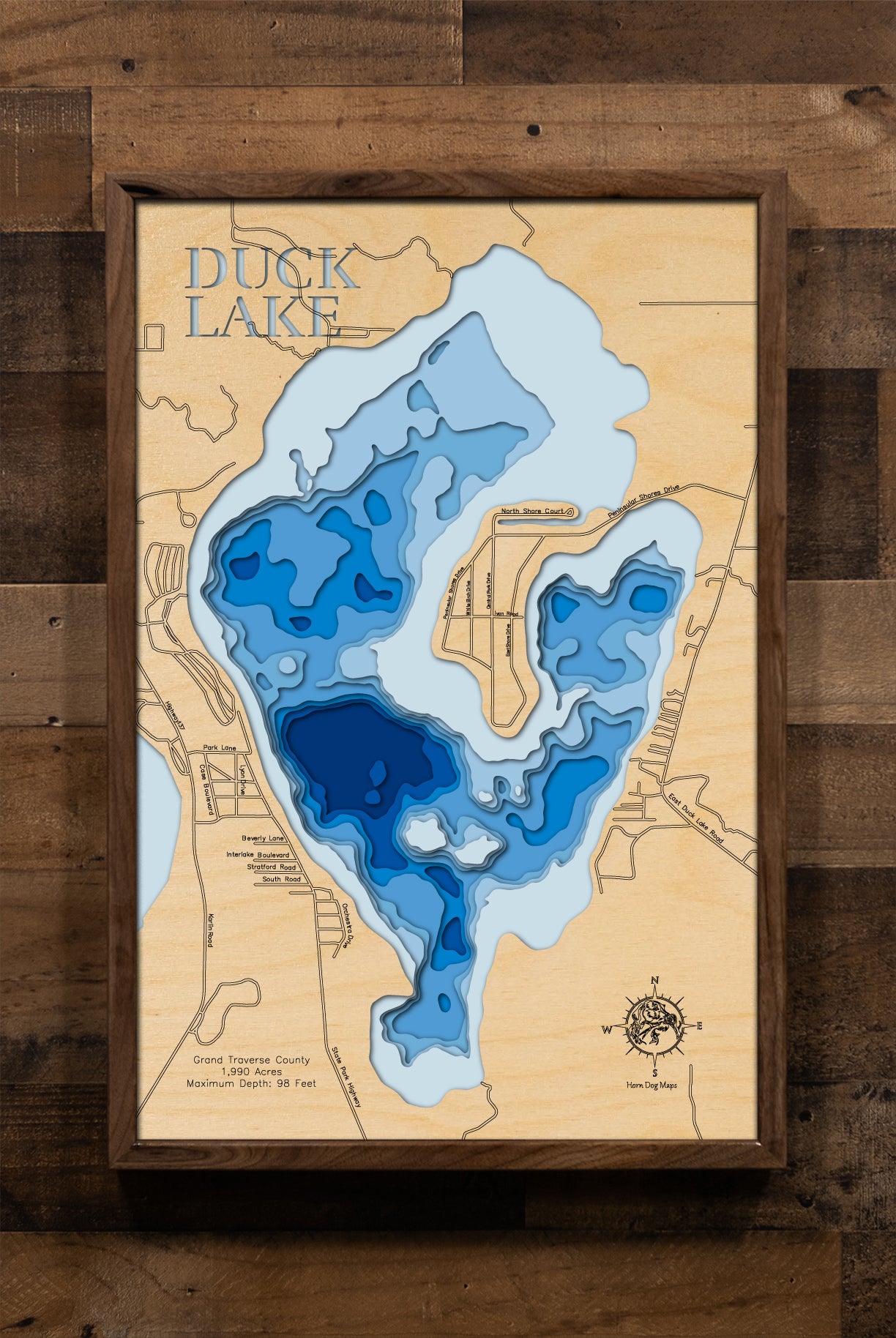 A laser-cut, layered wooden map of Duck Lake in Grand Traverse County, Michigan. The distinct shape of this 1,930-acre lake is visible within the northwestern Lower Michigan landscape. The varying depths of the lake, reaching a maximum of 98 feet and detailed by 10-foot contour intervals, are represented by different layers of wood, illustrating its underwater topography.