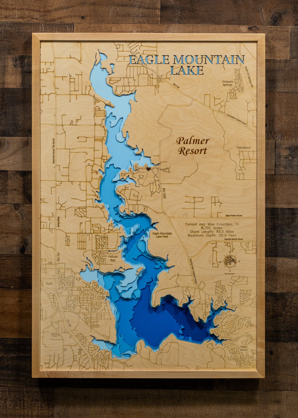 A framed 3D wooden depth map of Eagle Mountain Lake, featuring layered blue and light blue wood sections representing the lake, with engraved topography details and text including 'Eagle Mountain Lake' and 'Palmer Resort'. The frame is mounted on a rustic dark wood plank background.