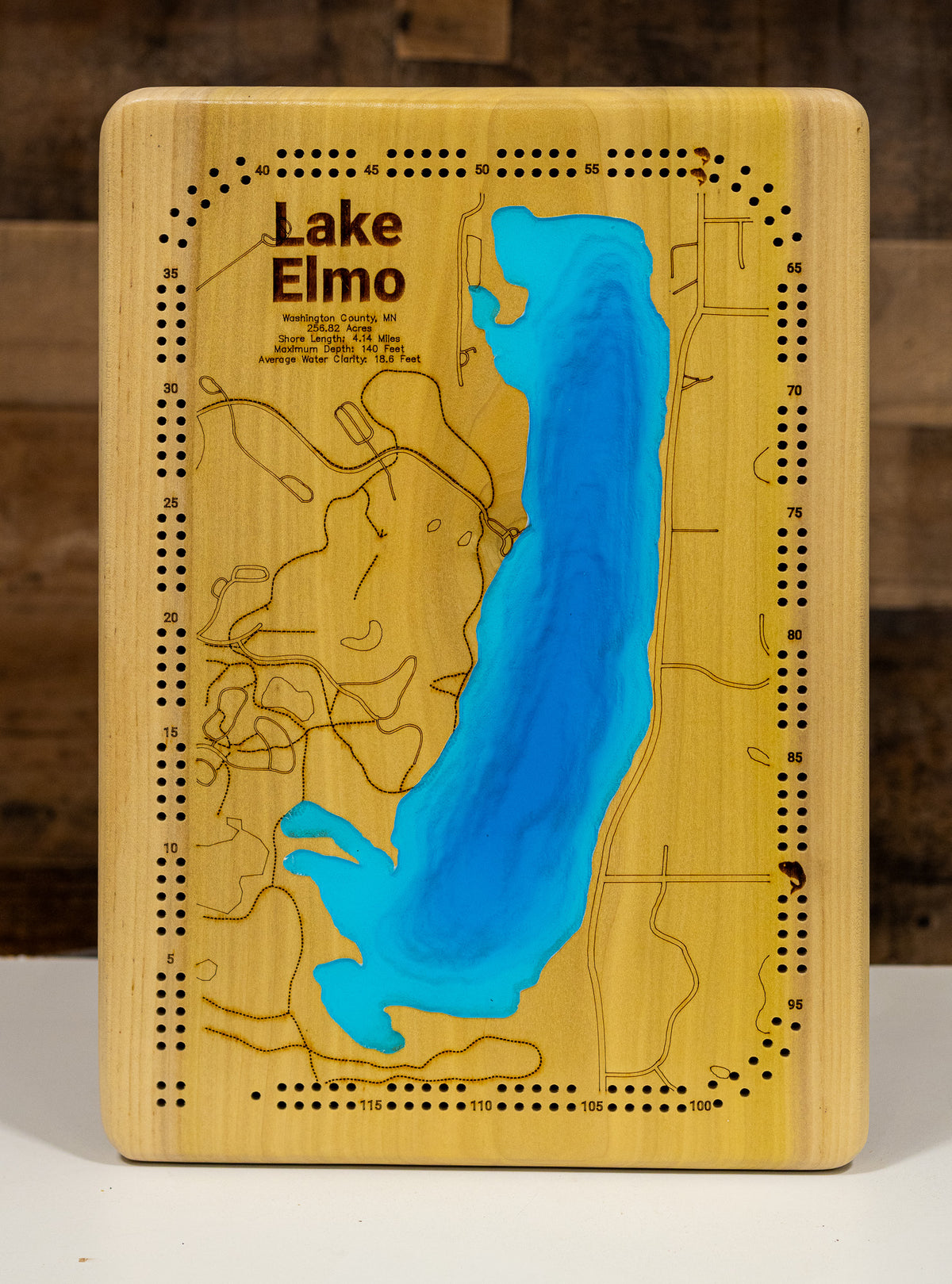 A highly detailed, laser-cut, and multi-layered wooden map of Lake Elmo, a natural lake in Washington County, Minnesota. The map shows the lake's distinct shape, its shoreline of 3.31 miles, and its central location within Lake Elmo Park Reserve. The varying depths of the lake, reaching a maximum of 44 feet, are represented by different layers of wood.