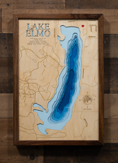 A highly detailed, laser-cut, and multi-layered wooden map of Lake Elmo, a natural lake in Washington County, Minnesota. The map shows the lake's distinct shape, its shoreline of 3.31 miles, and its central location within Lake Elmo Park Reserve. The varying depths of the lake, reaching a maximum of 44 feet, are represented by different layers of wood.