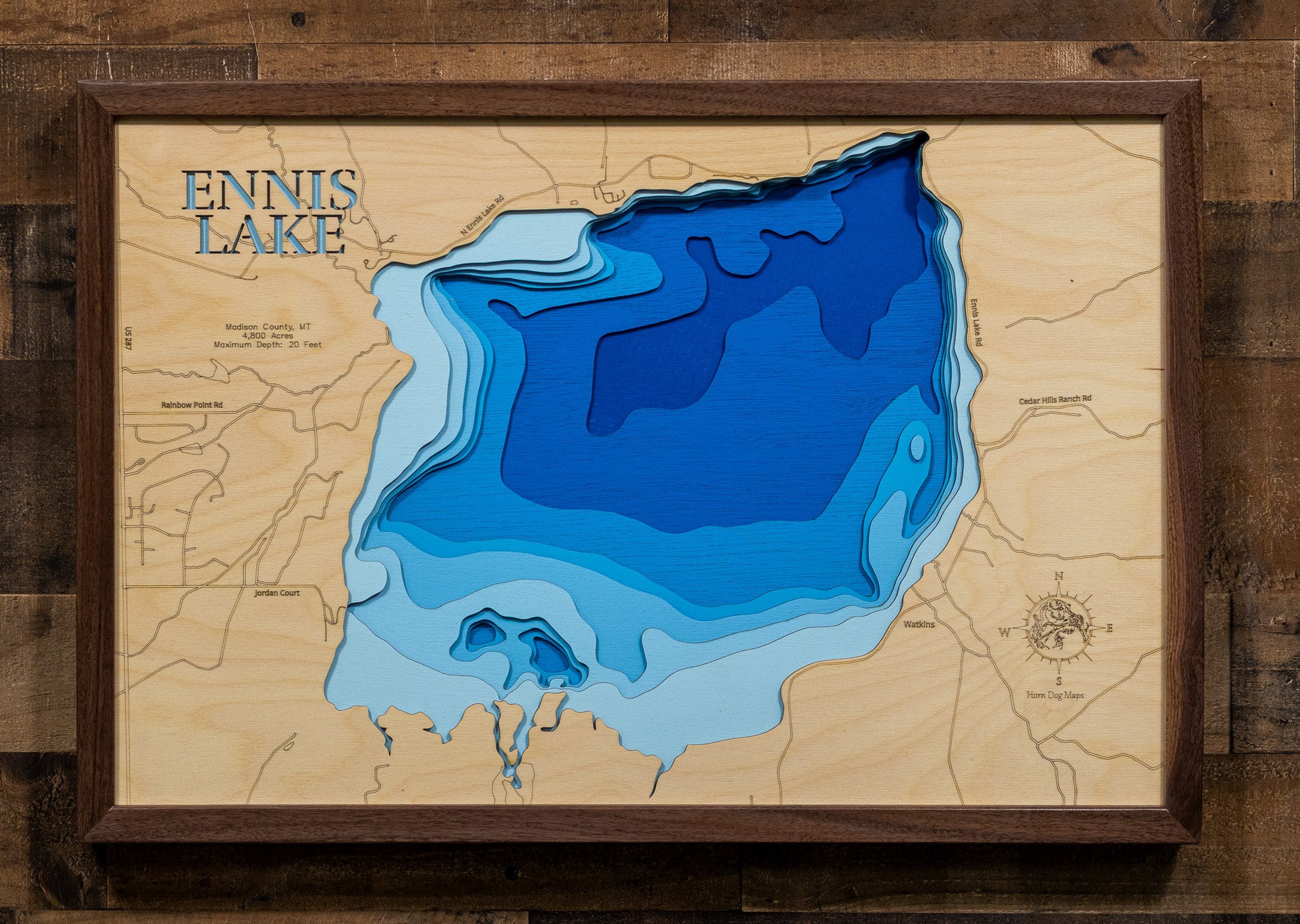 A highly detailed, laser-cut, and multi-layered wooden map of Ennis Lake, a reservoir in Madison County, Montana. The map shows the lake's distinct shape and its location on the Madison River. The varying depths of the lake, reaching a maximum of 20 feet, are represented by different layers of wood, illustrating its impressive underwater topography.