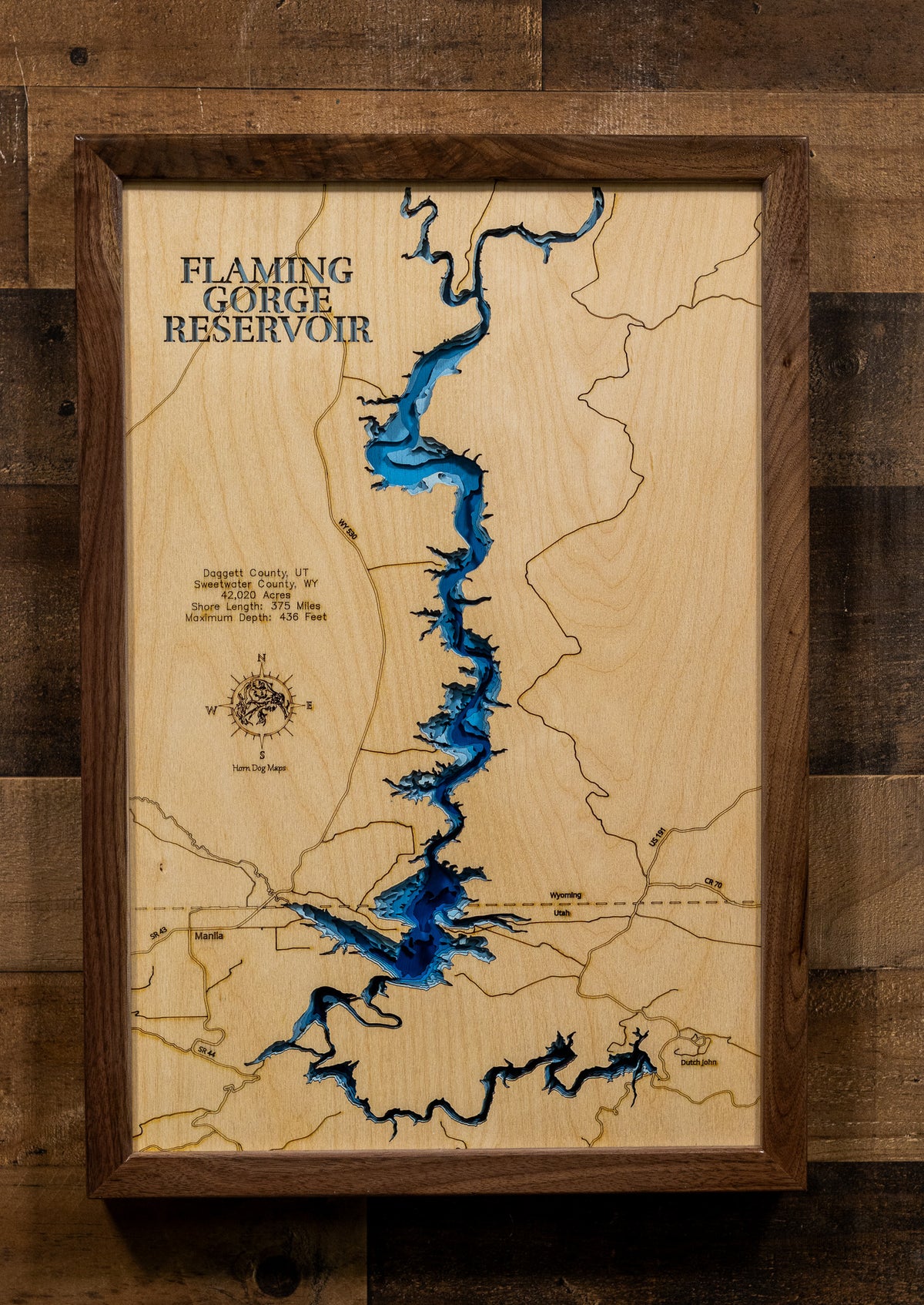 A framed wooden map of Flaming Gorge Reservoir, featuring a light natural wood background, dark brown engraved lines, and a vivid blue reservoir. The piece is housed in a dark brown wooden frame and mounted on a rustic dark wood plank wall.