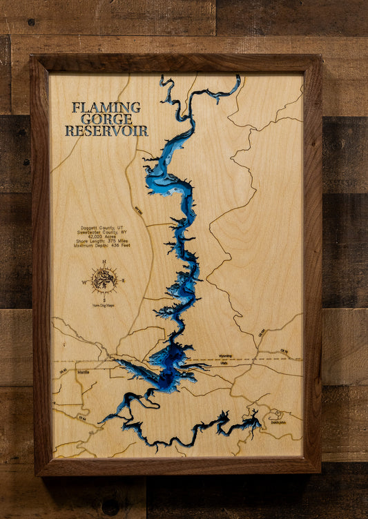 A framed wooden map of Flaming Gorge Reservoir, featuring a light natural wood background, dark brown engraved lines, and a vivid blue reservoir. The piece is housed in a dark brown wooden frame and mounted on a rustic dark wood plank wall.