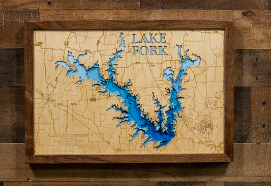 A framed wooden lake map wall art piece featuring a detailed cutout design of Lake Fork, with blue water layers and a warm natural wood frame, mounted on a rustic wood plank wall.