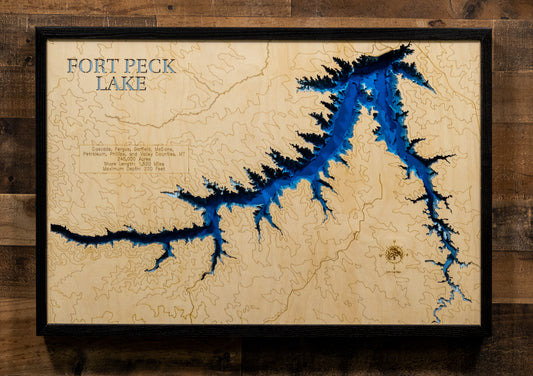 A framed Fort Peck Lake topographic map wall art, featuring deep blue water channels carved into a light natural wood background, mounted on a rustic dark wood plank wall.