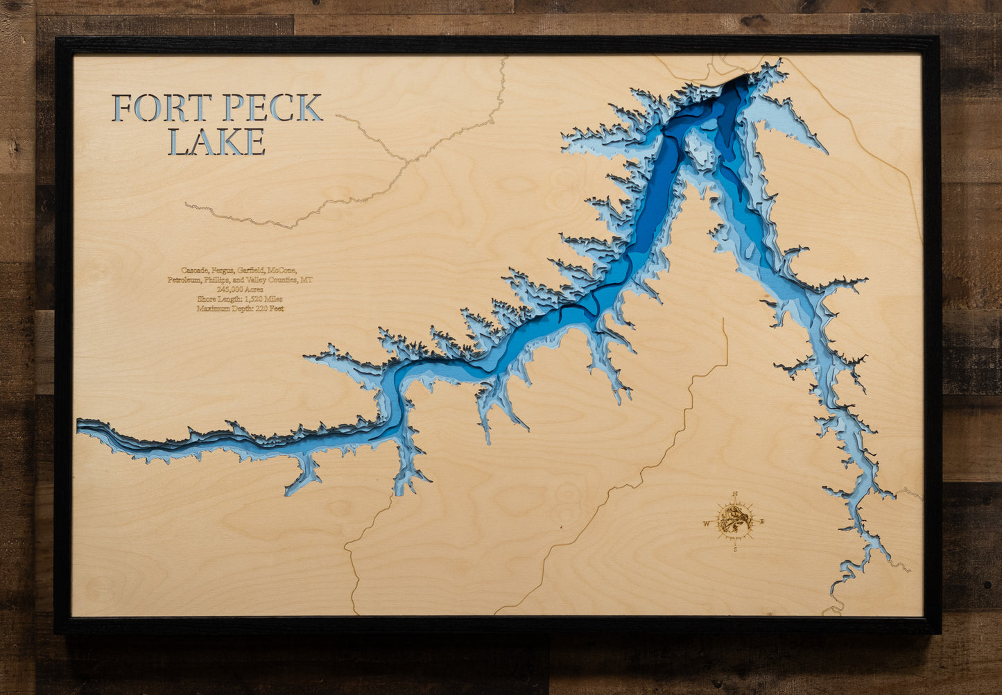 A laser-cut, layered wooden map of Fort Peck Lake in Montana. The expansive and intricate shape of this massive reservoir, winding through the landscape of northeastern Montana, is clearly visible. The varying depths of the lake, reaching a maximum of 220 feet, are represented by different layers of wood, illustrating its impressive underwater topography.
