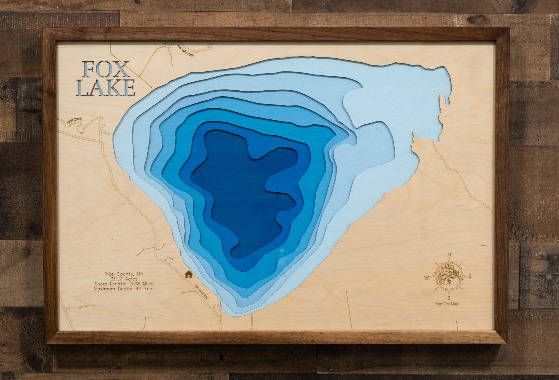 A laser-cut, layered wooden map of Fox Lake in Rice County, Minnesota. The distinct shape of this natural lake, with its 3.08-mile shoreline, is clearly visible within the landscape of Rice County. The varying depths of the lake, reaching a maximum of 47 feet, are represented by different layers of wood, illustrating its impressive underwater topography.