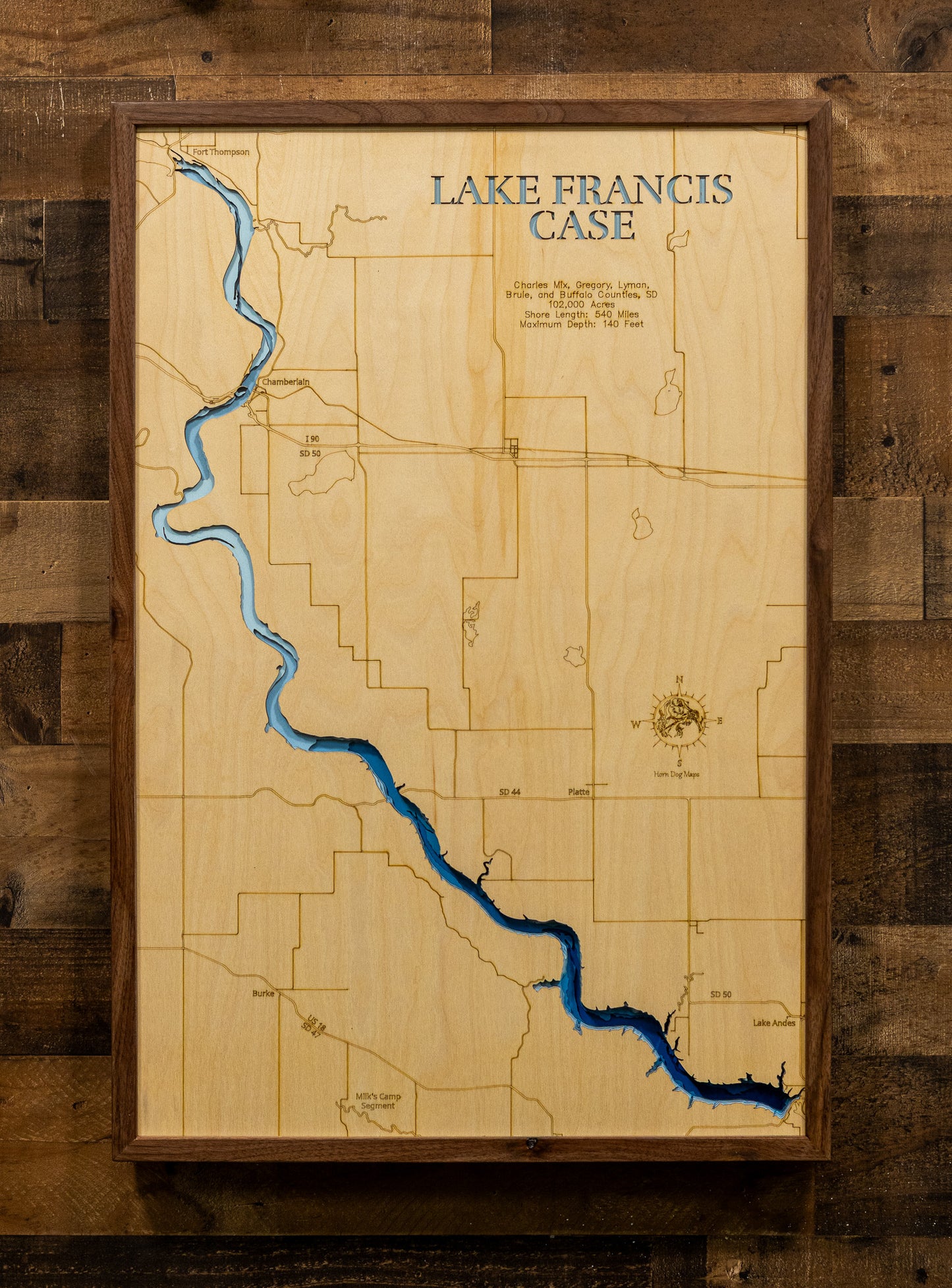 A framed wooden map of Lake Francis Case, featuring a light natural wood background, engraved county lines, and a winding blue lake representation, mounted on a rustic dark wood plank wall.