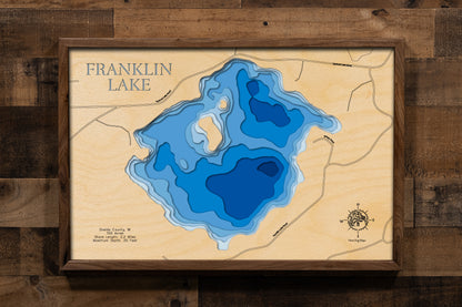 A laser-cut, layered wooden map of Franklin Lake in Oneida County, Wisconsin. The distinct shape of this natural lake, with its 3.53-mile shoreline, is clearly visible within the forested landscape near Minocqua in northern Wisconsin. The varying depths of the lake, reaching a maximum of 61 feet, are represented by different layers of wood, illustrating its impressive underwater topography.