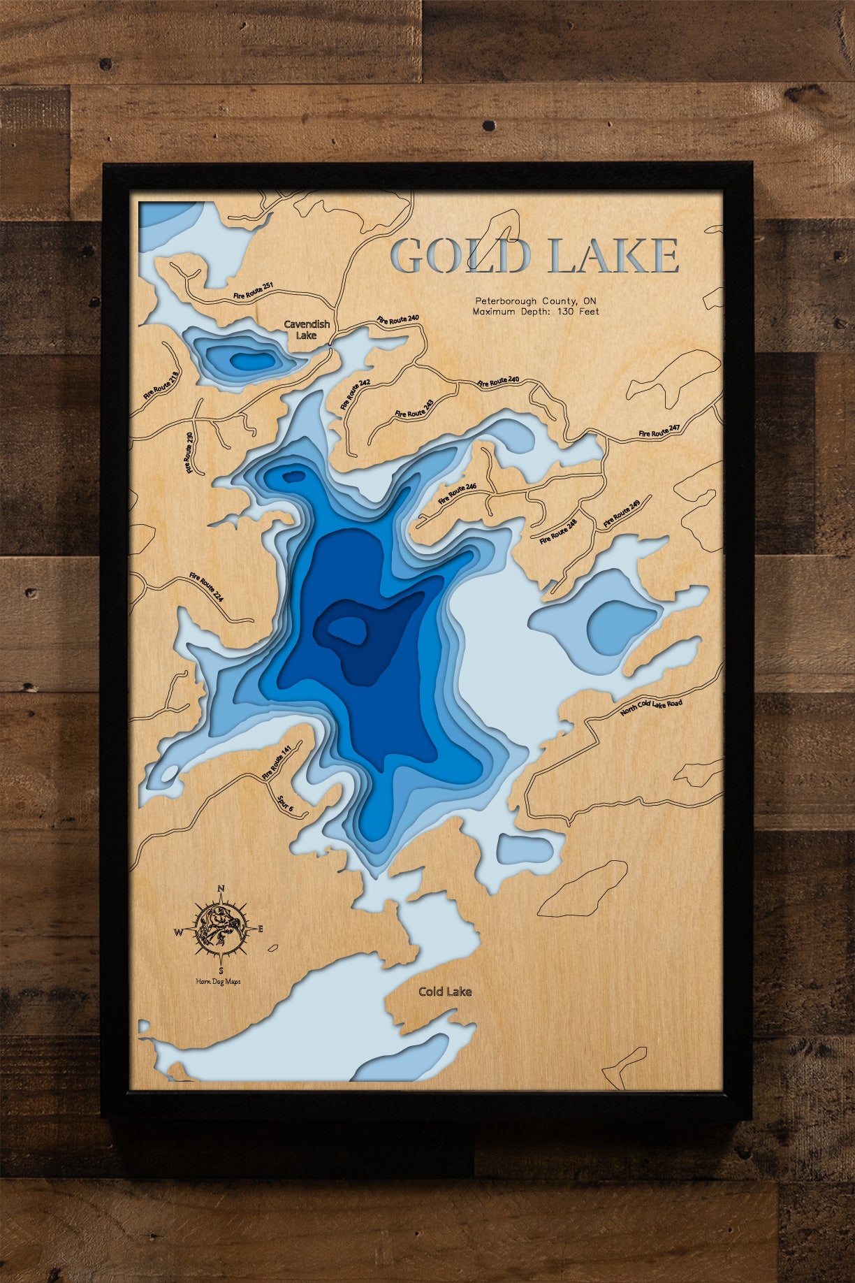 A precision laser-cut, multi-layered wooden map of Gold Lake in Peterborough County, Ontario — a deep Canadian Shield lake reaching 130 feet in the Municipality of Trent Lakes near Kawartha Highlands Provincial Park. The natural wood top layer traces the lake's dramatic, irregular shoreline in fine detail, while a light-to-dark blue gradient conveys its striking depth. A handcrafted statement piece for any Ontario cottage, Shield cabin, or Kawartha Lakes enthusiast's wall.