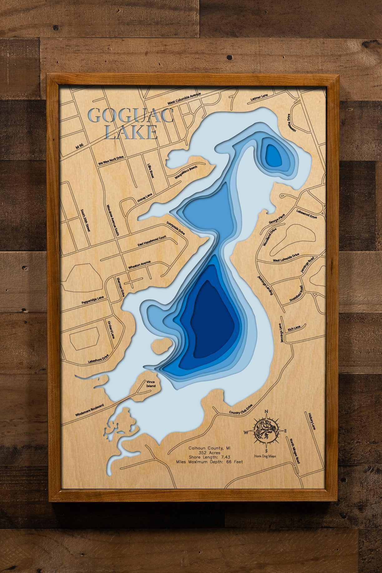 A highly detailed, laser-cut, and multi-layered wooden map of Goguac Lake, a natural lake in Calhoun County, Michigan. The map shows the lake's distinct shape, its 7.43-mile shoreline, and the three small islands. The varying depths of the lake, reaching a maximum of 66 feet, are represented by different layers of wood, illustrating its impressive underwater topography.