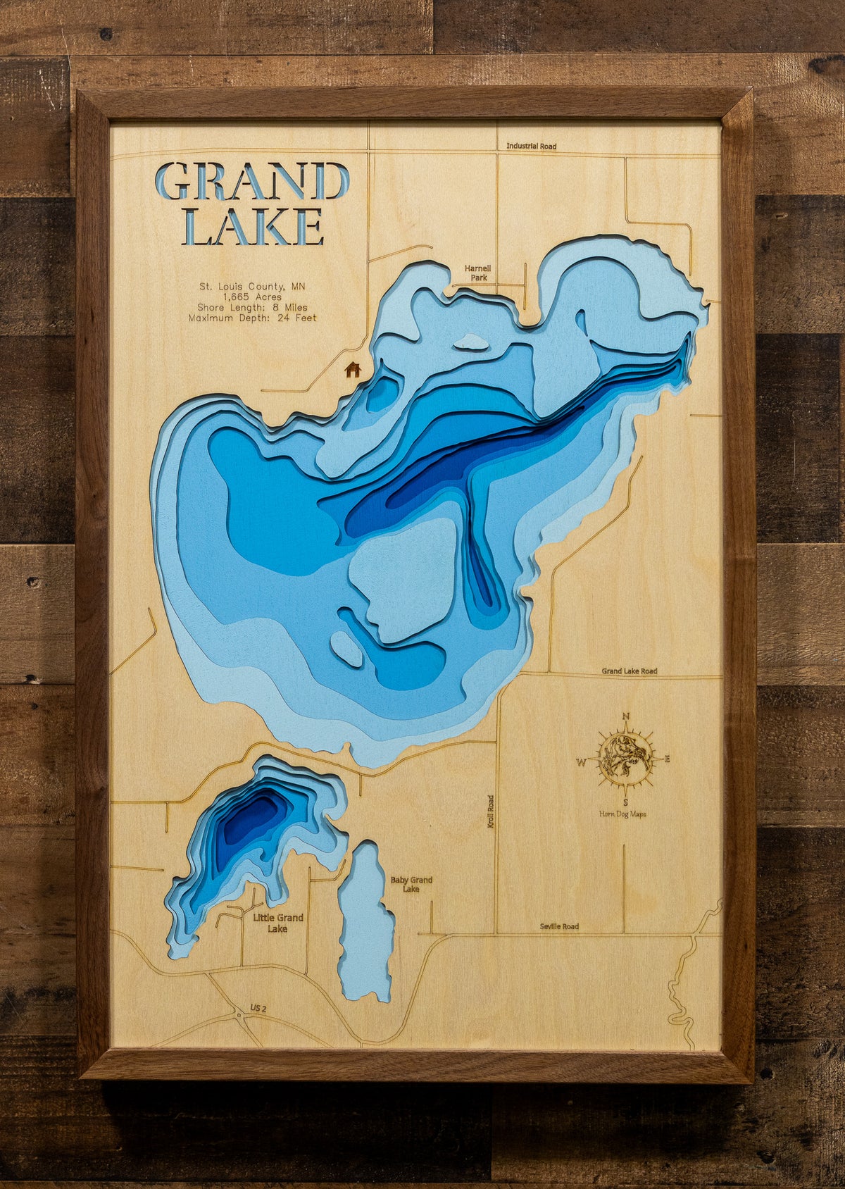 A framed wooden depth map of Grand Lake, featuring layered blue contours on a light natural wood background, mounted on a dark rustic wood wall.