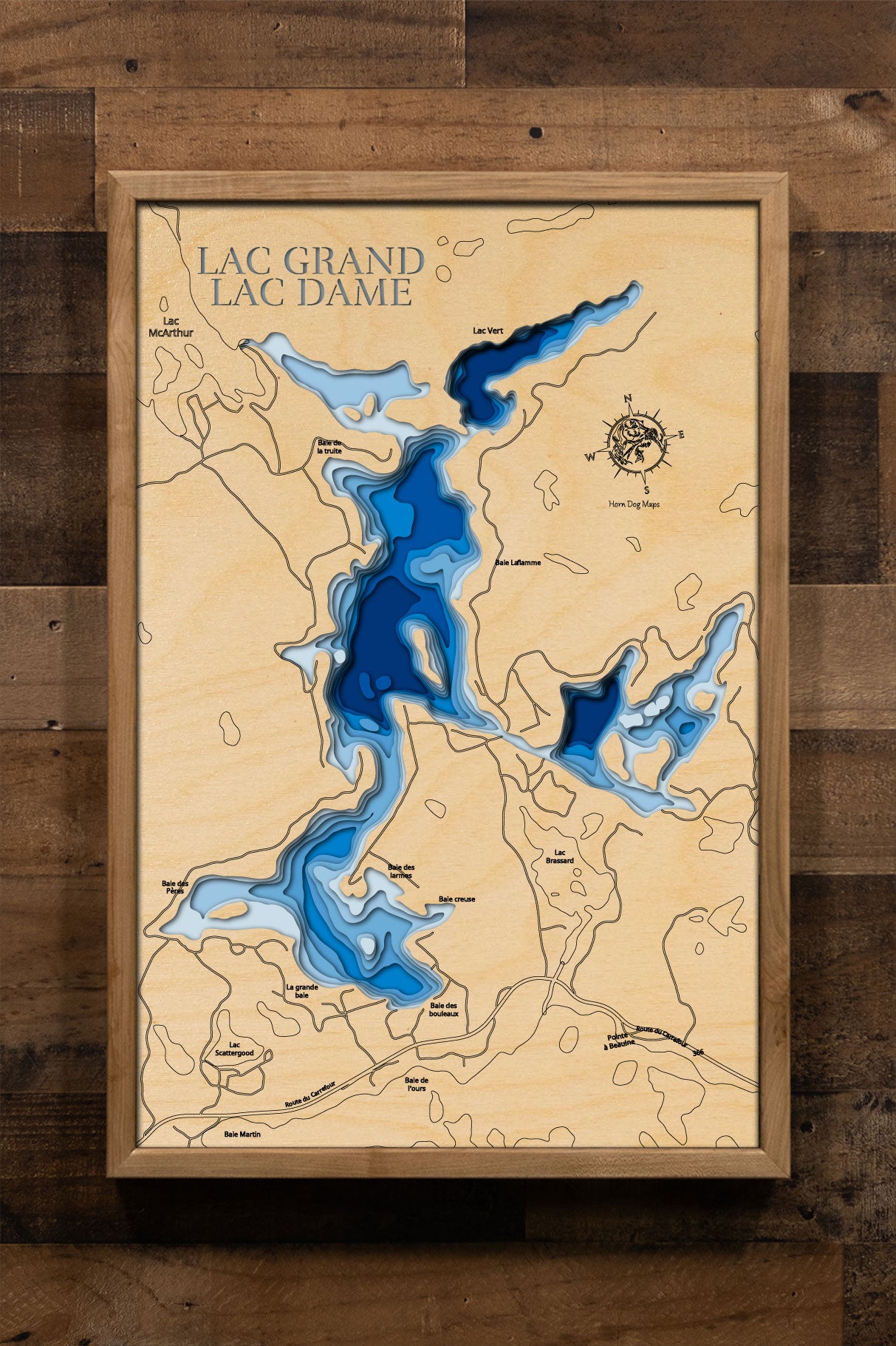 A laser-cut, layered wooden map of Lac Dame and Lac Grand in Val-des-Monts, Quebec. The interconnected shapes of the two lakes are visible within the hilly landscape of the region. The varying depths of Lac Grand, reaching 130 feet, are represented by different layers of wood, while the shallower and less documented depths of Lac Dame are also indicated.
