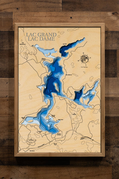 A laser-cut, layered wooden map of Lac Dame and Lac Grand in Val-des-Monts, Quebec. The interconnected shapes of the two lakes are visible within the hilly landscape of the region. The varying depths of Lac Grand, reaching 130 feet, are represented by different layers of wood, while the shallower and less documented depths of Lac Dame are also indicated.
