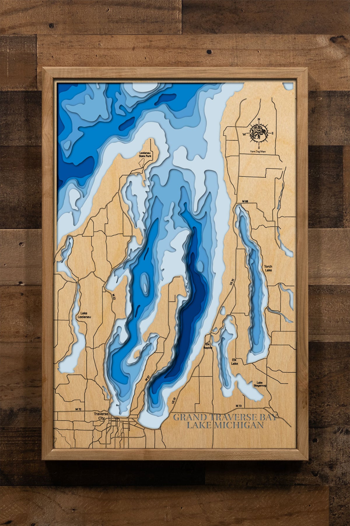 A precision laser-cut, multi-layered wooden map of Grand Traverse Bay in northern Michigan, a 177,280-acre bay of Lake Michigan divided by the Old Mission Peninsula into East and West arms with 132 miles of shoreline and a maximum depth of 590 feet. The natural wood top layer captures every dramatic curve of this iconic bay in fine detail, while a light-to-dark blue gradient conveys its remarkable depth and crystal-clear character. A handcrafted statement piece for any northern Michigan home, Traverse City 