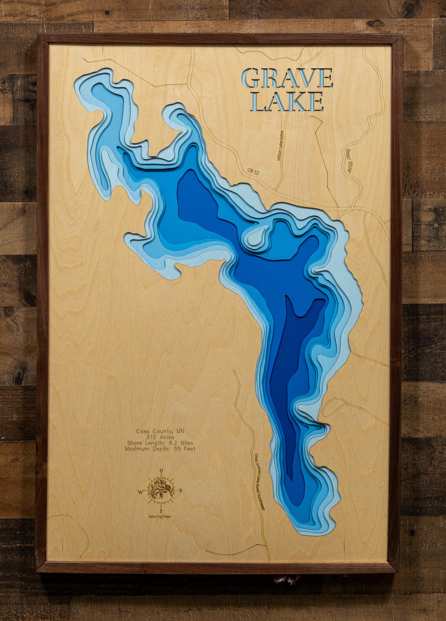 A meticulously detailed, laser-cut, multi-layered wooden topographical map of Grave Lake in Cass County, Minnesota, showcasing the 372-acre lake's irregularly shaped surface with numerous bays and points along 6.2 miles of intricate shoreline. The natural wood top layer frames the exceptionally clear Lake Class 23 water with depth contours from shallow littoral areas to the 55-foot maximum depth, while graduated blue layers create a striking visual depth gradient.