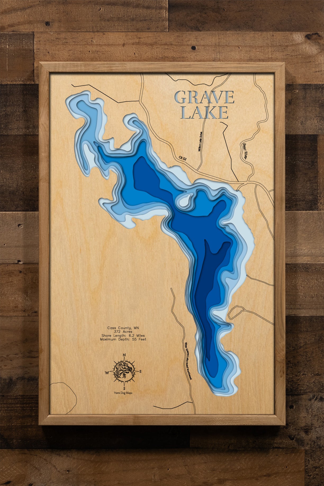 A meticulously detailed, laser-cut, multi-layered wooden topographical map of Grave Lake in Cass County, Minnesota, showcasing the 372-acre lake's irregularly shaped surface with numerous bays and points along 6.2 miles of intricate shoreline. The natural wood top layer frames the exceptionally clear Lake Class 23 water with depth contours from shallow littoral areas to the 55-foot maximum depth, while graduated blue layers create a striking visual depth gradient.