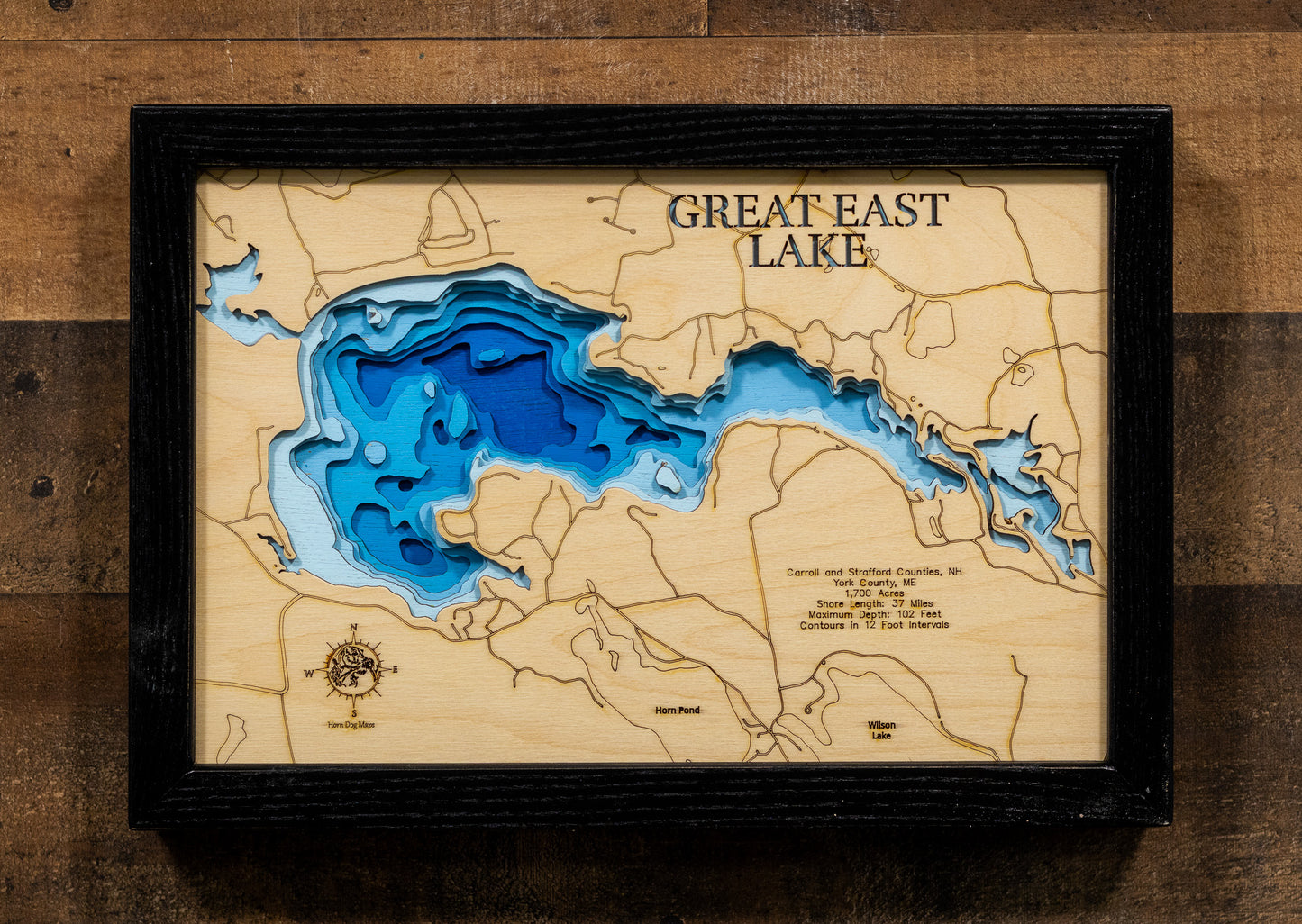 A laser-cut, layered wooden map of Great East Lake in New Hampshire and Maine. The distinctive and intricate shape of this large border lake, with its numerous coves and islands along its 37-mile shoreline, is clearly visible across the landscape shared by Carroll and Strafford Counties, NH, and York County, ME. The varying depths of the lake, reaching a maximum of 102 feet and detailed by 12-foot contour intervals, are represented by different layers of wood, illustrating its impressive underwater topograp