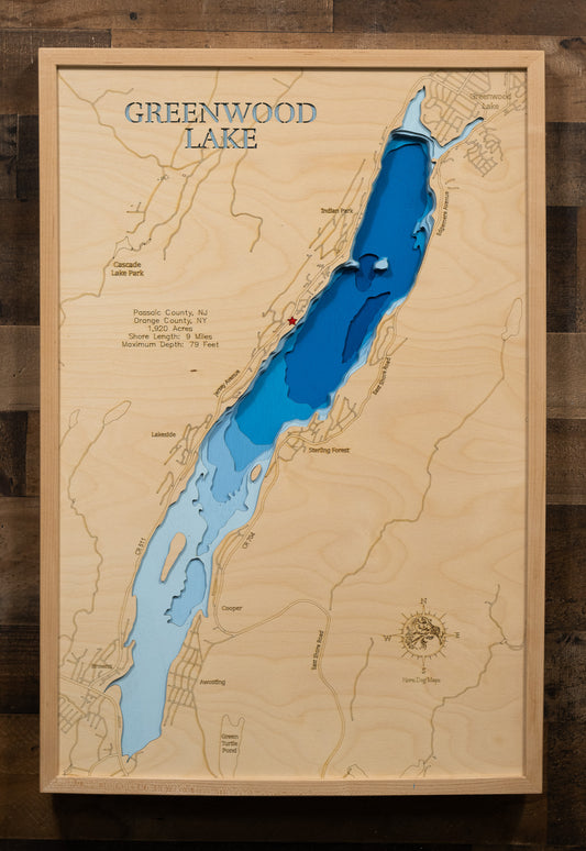 A laser-cut, layered wooden map of Greenwood Lake in New Jersey and New York. The distinct, elongated shape of this interstate lake, with its 9-mile shoreline, is clearly visible as it stretches across the landscape of Passaic County, NJ, and Orange County, NY. The varying depths of the lake, reaching a maximum of 79 feet, are represented by different layers of wood, illustrating its underwater topography.