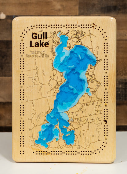 A cribbage board featuring an engraved map of Gull Lake with blue resin-filled inlays, photographed against a rustic wood background.