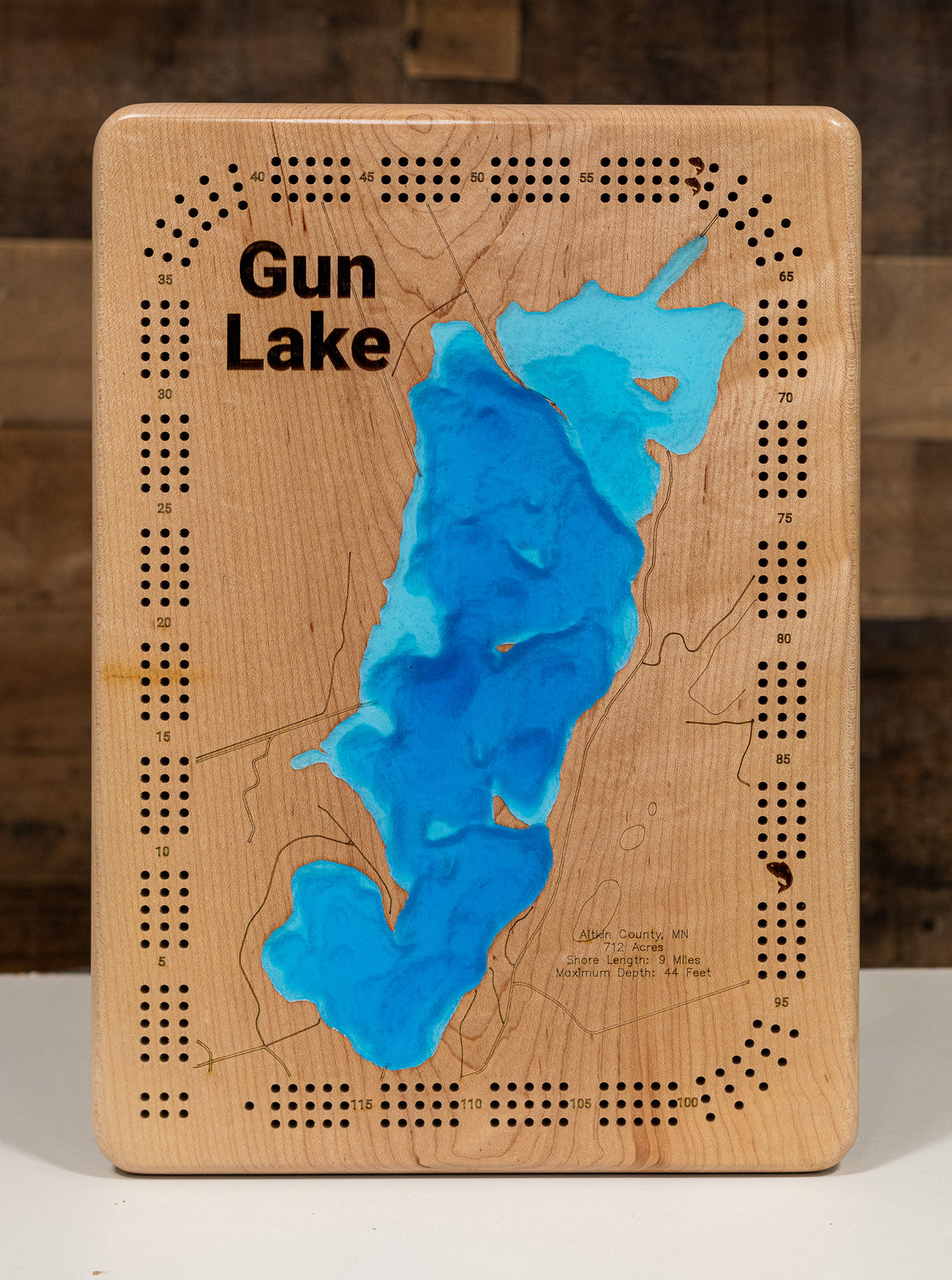 A Cribbage board featuring a topographic-style map of Gun Lake with blue resin-filled depressions and black engraved text and dotted border details, photographed against a rustic wood background.