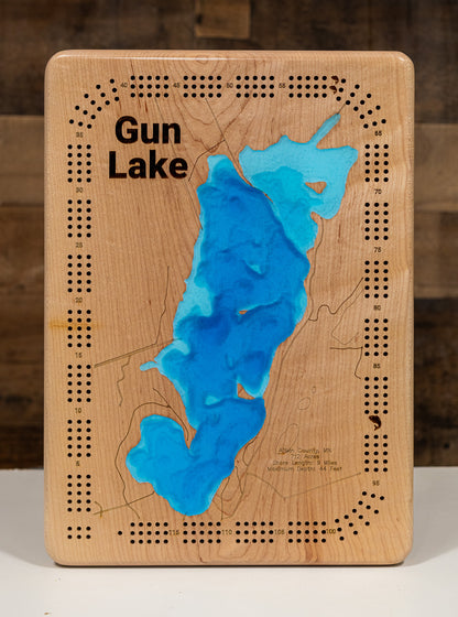 A Cribbage board featuring a topographic-style map of Gun Lake with blue resin-filled depressions and black engraved text and dotted border details, photographed against a rustic wood background.