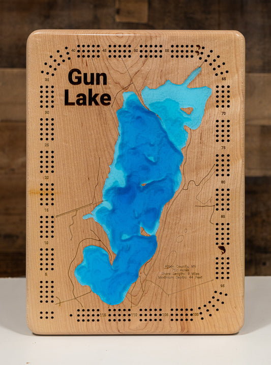 A Cribbage board featuring a topographic-style map of Gun Lake with blue resin-filled depressions and black engraved text and dotted border details, photographed against a rustic wood background.