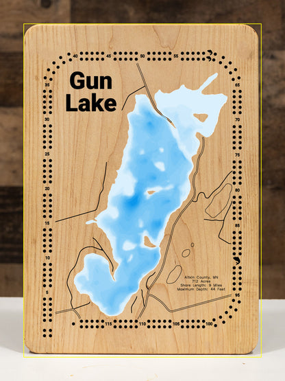 A meticulously detailed, laser-cut, multi-layered wooden topographical map of Gun Lake in Aitkin County, Minnesota, showcasing the 712-acre surface with intricate depth contours from diverse vegetated areas to the 44-foot maximum depth. The natural wood top layer frames nine miles of shoreline with abundant aquatic vegetation and the public boat access, while graduated blue layers create a striking visual depth gradient.