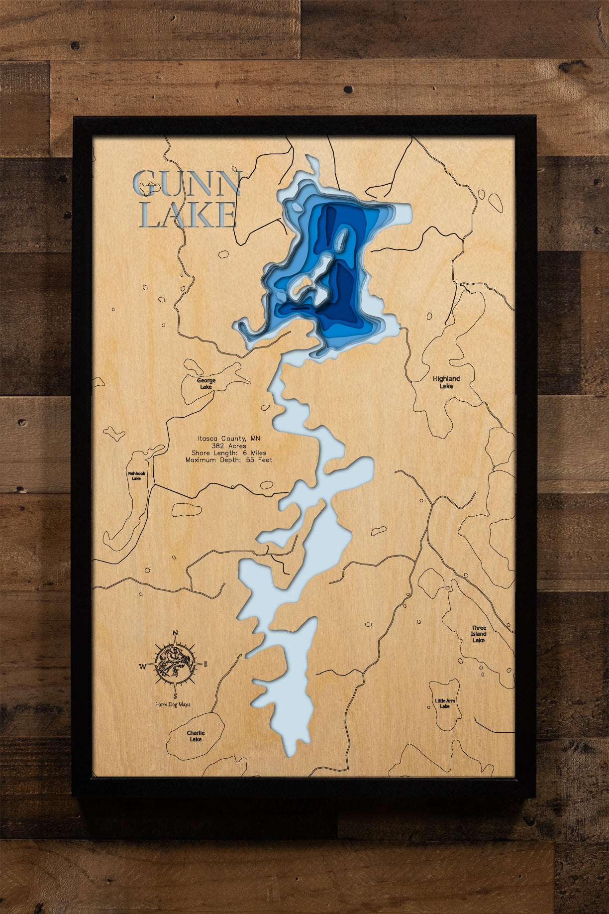 A meticulously detailed, laser-cut, multi-layered wooden topographical map of Gunn Lake in Itasca County, Minnesota, showcasing the 382-acre lake's series of connected basins with depth contours from the 17-foot average to the 55-foot maximum depth and exceptional 12.3-foot water clarity. The natural wood top layer frames approximately six miles of lightly developed shoreline, inlets from Charlie and Highland Lakes, the outlet to Fox Lake, and the state-owned public access, while graduated blue layers creat
