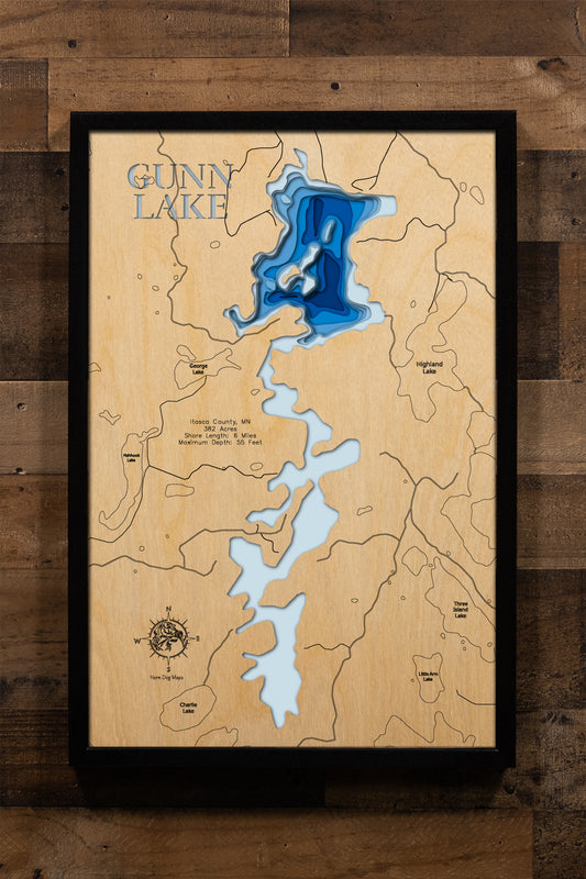 A meticulously detailed, laser-cut, multi-layered wooden topographical map of Gunn Lake in Itasca County, Minnesota, showcasing the 382-acre lake's series of connected basins with depth contours from the 17-foot average to the 55-foot maximum depth and exceptional 12.3-foot water clarity. The natural wood top layer frames approximately six miles of lightly developed shoreline, inlets from Charlie and Highland Lakes, the outlet to Fox Lake, and the state-owned public access, while graduated blue layers creat