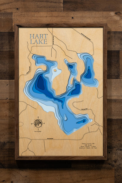 A meticulously detailed, laser-cut, multi-layered wooden topographical map of Hart Lake in Itasca County, Minnesota, showcasing the lake's 328-acre surface with intricate depth contours illustrating wetland inlets and the transition from shallow areas to the 55-foot maximum depth. The natural wood top layer frames seven miles of precisely rendered shoreline and the outlet to Swan Lake, while graduated blue layers create a striking visual depth gradient.