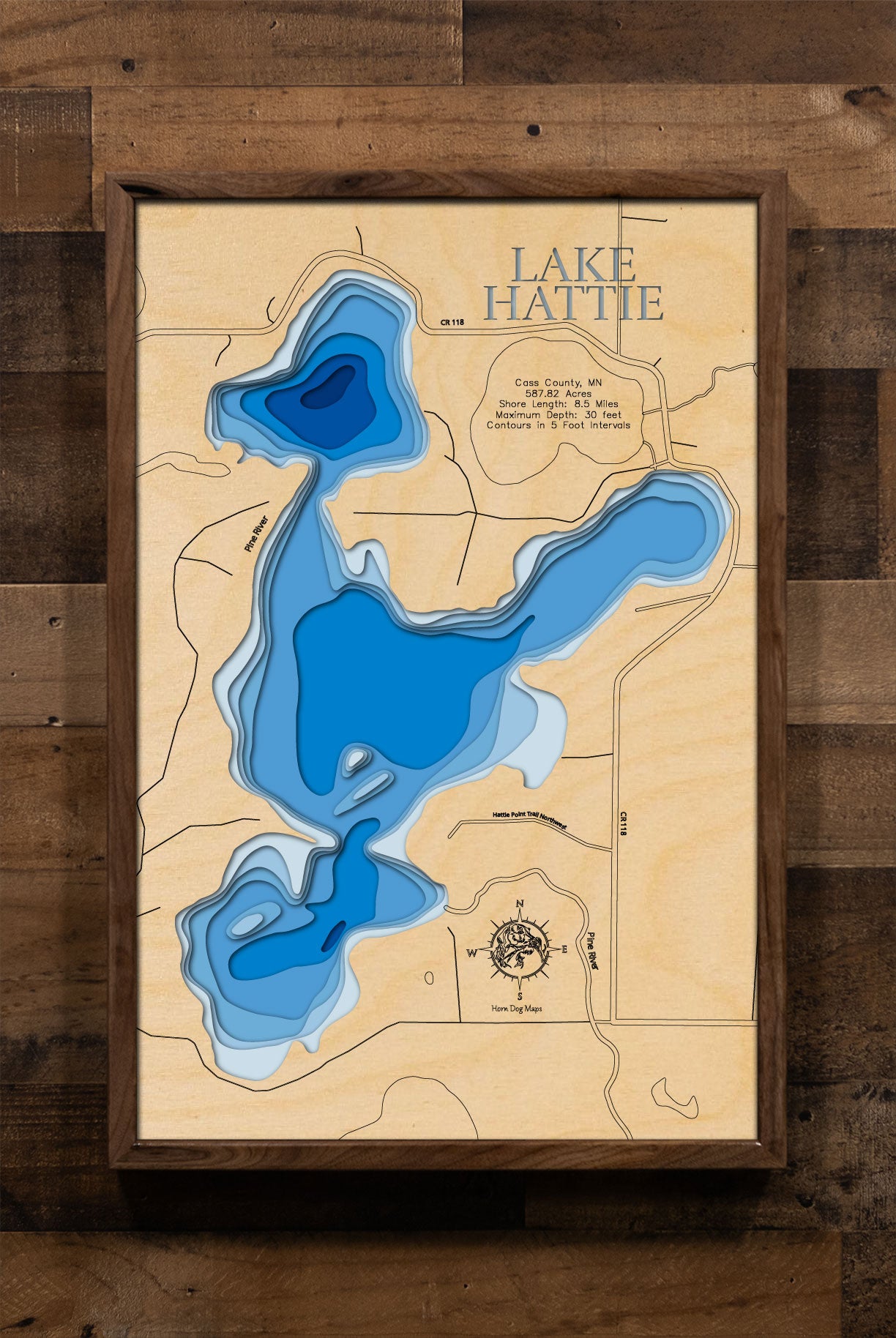 A meticulously crafted laser-cut wooden lake map of Lake Hattie in Cass County, Minnesota. The detailed map features accurate, layered topography with intricate, engraved 5-foot contour lines. The water sections are stained a rich blue, providing a striking contrast to the natural wood of the landmasses and surrounding shoreline. The map includes engraved labels for the lake name, county, and state, and highlights key data points such as maximum depth and shore length, capturing the unique geography of this