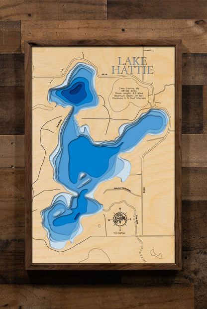 A meticulously crafted laser-cut wooden lake map of Lake Hattie in Cass County, Minnesota. The detailed map features accurate, layered topography with intricate, engraved 5-foot contour lines. The water sections are stained a rich blue, providing a striking contrast to the natural wood of the landmasses and surrounding shoreline. The map includes engraved labels for the lake name, county, and state, and highlights key data points such as maximum depth and shore length, capturing the unique geography of this