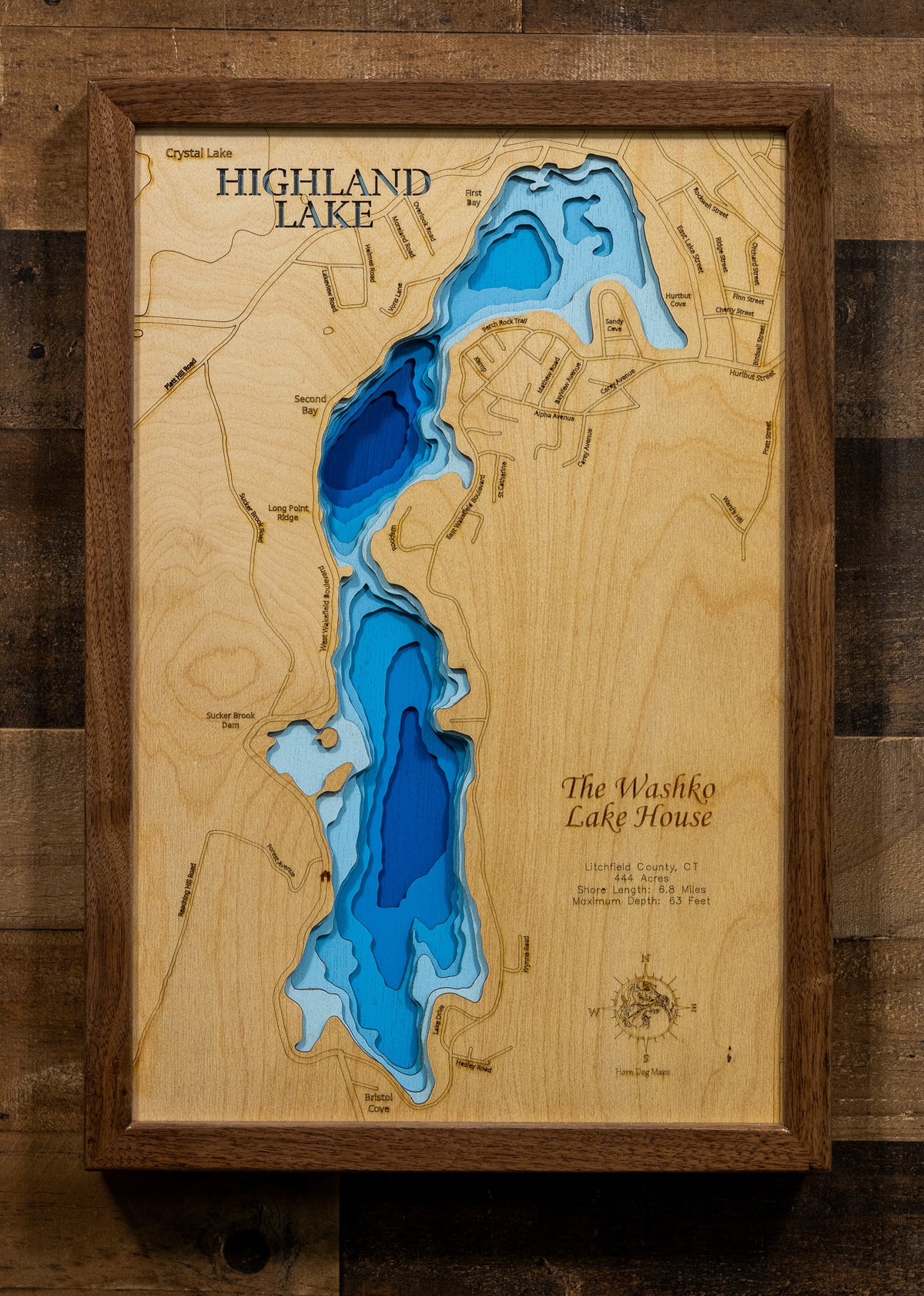 A framed wooden topographical map of Highland Lake, featuring layered blue and white wood sections representing water depth on a natural light wood background, mounted on a dark rustic wood wall.