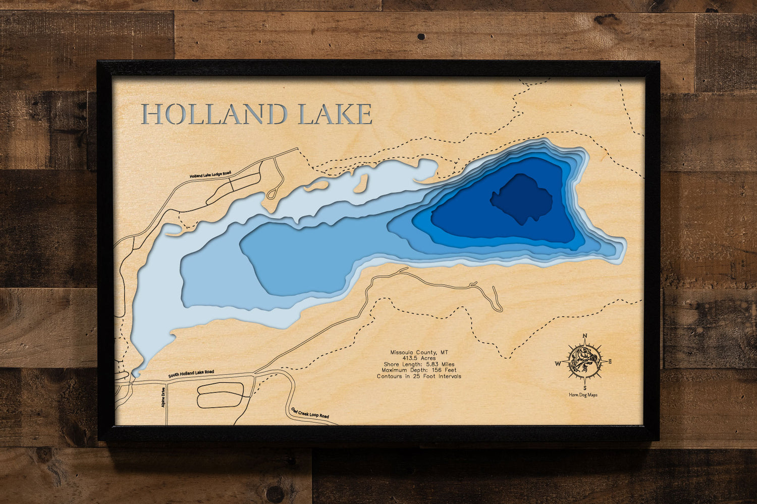 A laser-cut, layered wooden map of Holland Lake in Missoula County, Montana. The distinct shape of this natural lake, with its 5.83-mile shoreline, is clearly visible as it sits at the base of the Swan Range. The varying depths of the lake, reaching a maximum of 156 feet and detailed by 25-foot contour intervals, are represented by different layers of wood, illustrating its impressive underwater topography.