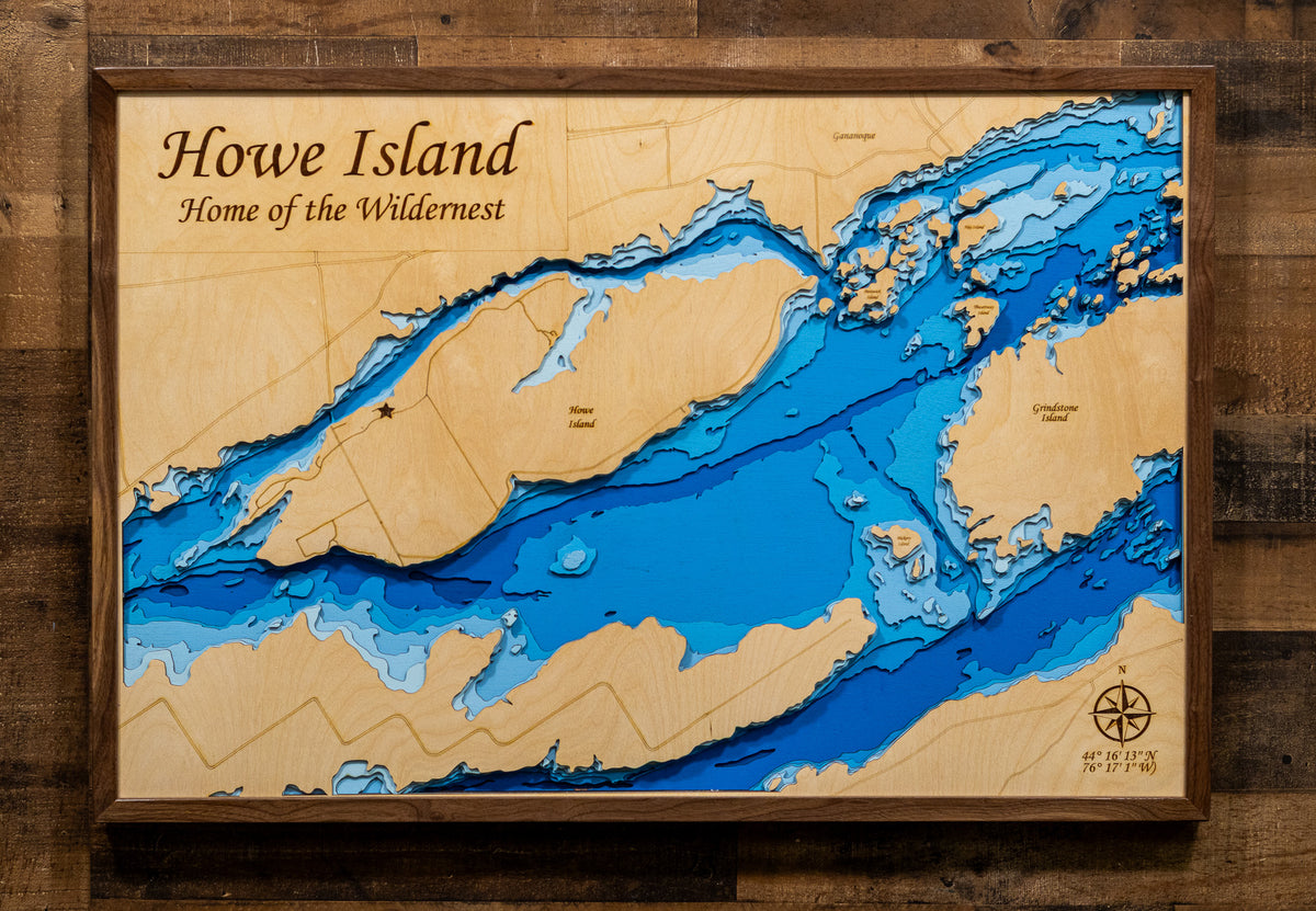 A framed wooden map of Howe Island, on the St Lawrence River, featuring layered blue water depth contours on a light wood background, mounted on a dark wood wall.
