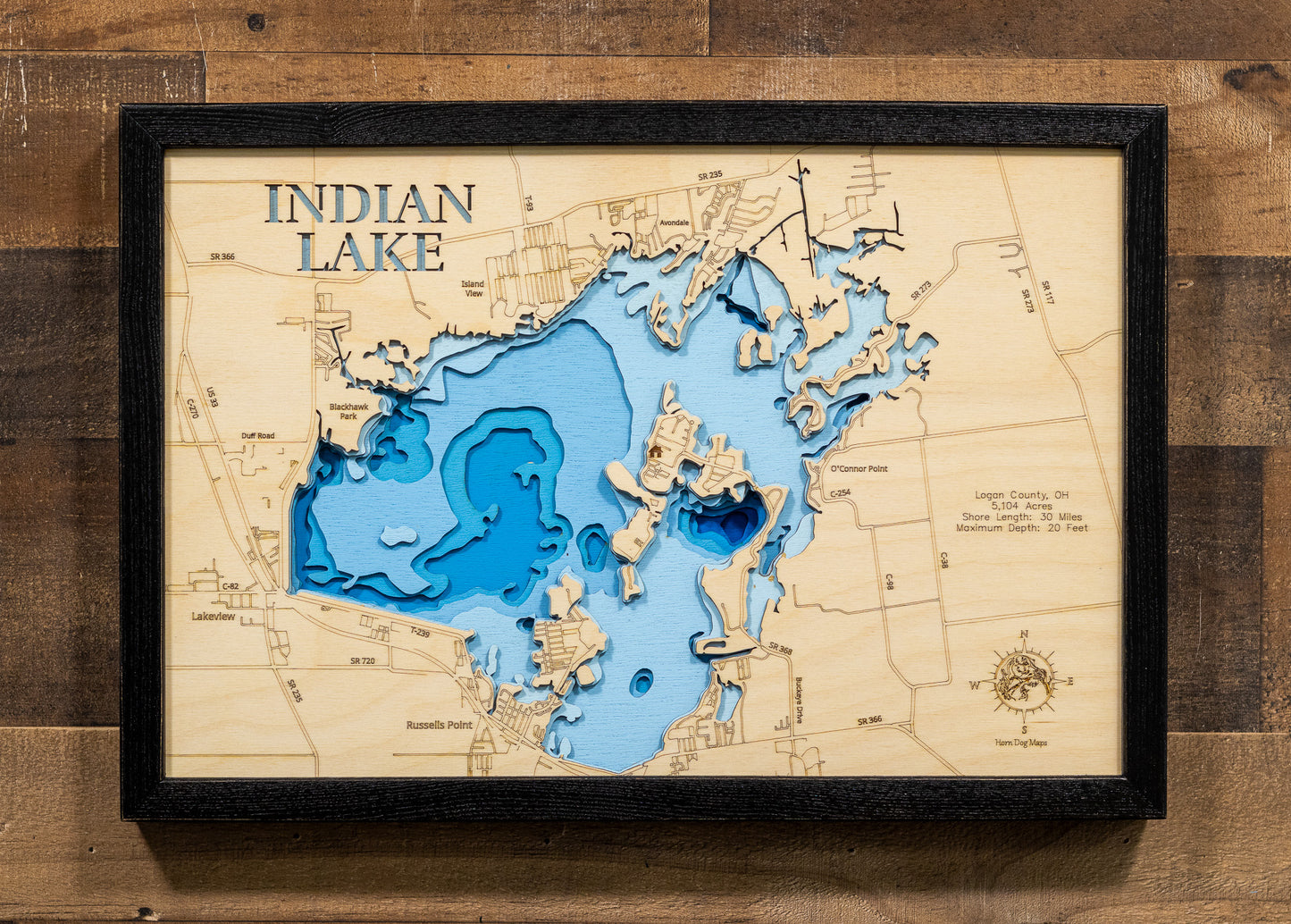 A highly detailed, laser-cut, and multi-layered wooden map of Indian Lake in Logan County, Ohio. The map showcases the reservoir's distinctive 5,104-acre shape with the iconic Indian Isles island chain arcing through its center, depth contours illustrating the shallow 20-foot maximum depth, and the intricate 30 miles of shoreline with numerous bays and inlets.